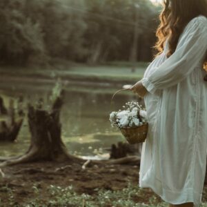 Pregnant woman in a white dress holds a basket of flowers by a peaceful river setting.